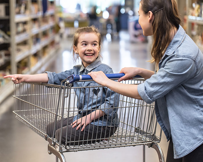 Mother pushing happy daughter in shopping cart in store aisle, capturing embarrassing moments parents will never forget thanks to kids