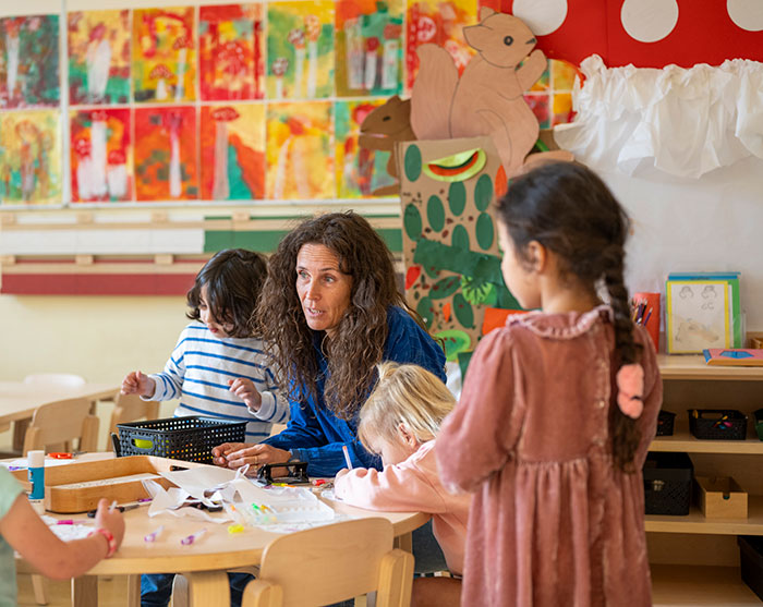 Teacher and young children engaged in arts and crafts in a colorful classroom with educational decorations and drawings.