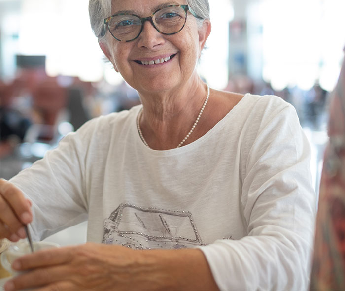 Smiling elderly woman wearing glasses and a white shirt, enjoying a casual moment in a bright indoor setting.