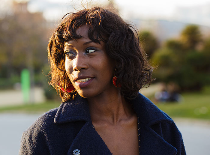 Young Black woman wearing a navy coat outdoors, looking to the side with a thoughtful expression in natural light.