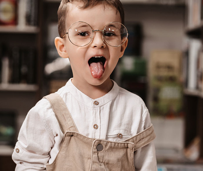 Young boy wearing glasses and beige overalls sticking out tongue in a playful embarrassing moments parents will never forget scene