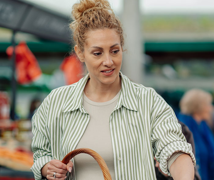 Woman with curly hair holding a basket at an outdoor market, reflecting on embarrassing moments parents experience with kids.