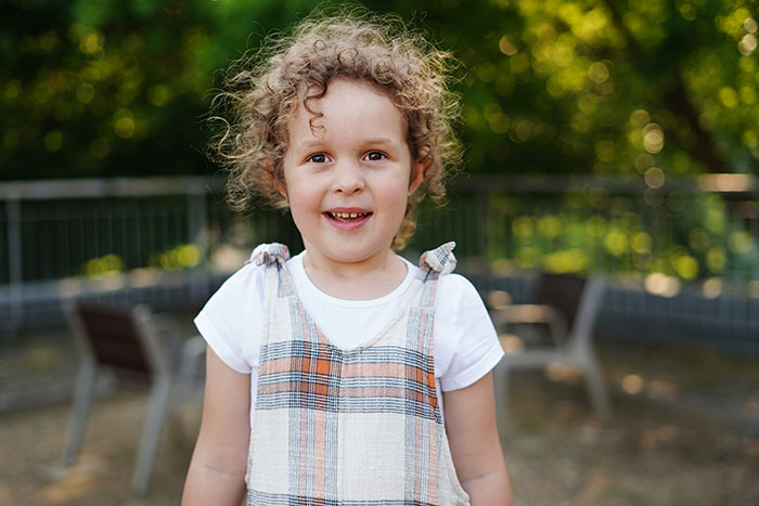 Smiling young child with curly hair wearing a plaid dress outdoors, capturing embarrassing moments parents will never forget.