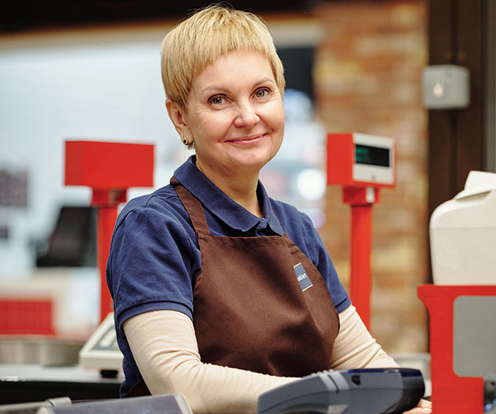 Smiling woman cashier wearing brown apron, standing behind counter with payment terminal, representing embarrassing moments parents.