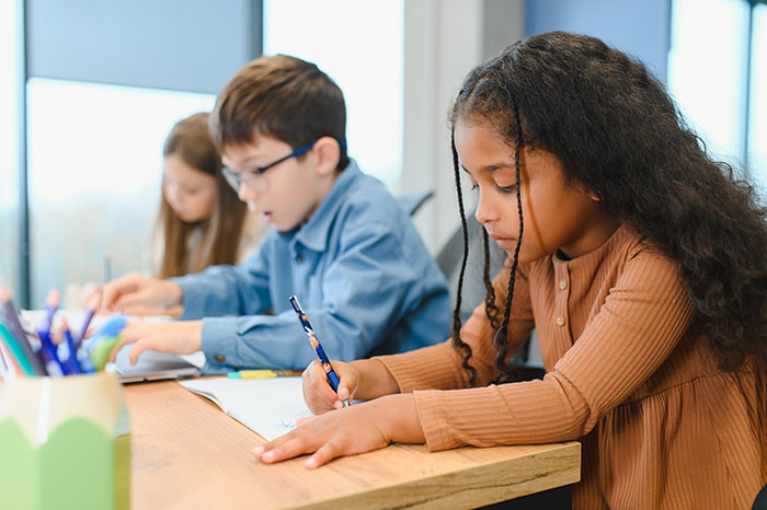 Three diverse kids concentrating on writing at a table, illustrating awkward and embarrassing moments parents experience with their kids.