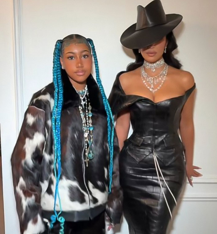 Kim Kardashian wearing a black corset dress and hat next to her daughter North West with long blue braids and a patterned jacket