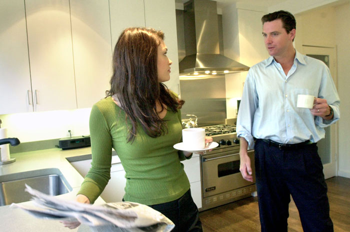  Kimberly Guilfoyle in green top holding a cup and newspaper talking to Gavin Newsom holding a coffee in a modern kitchen.