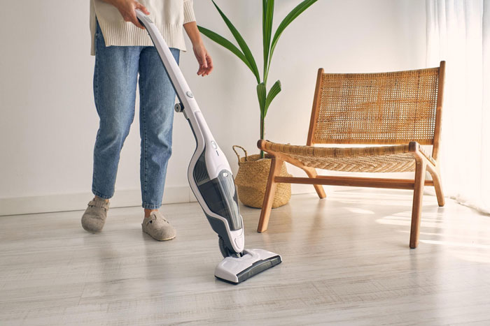 Person using a modern vacuum cleaner on light wood floor near wooden chair and potted plant in bright room.