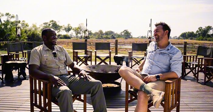 Two men enjoying a relaxed conversation on a wooden deck with the Okavango Delta's scenic beauty in the background.