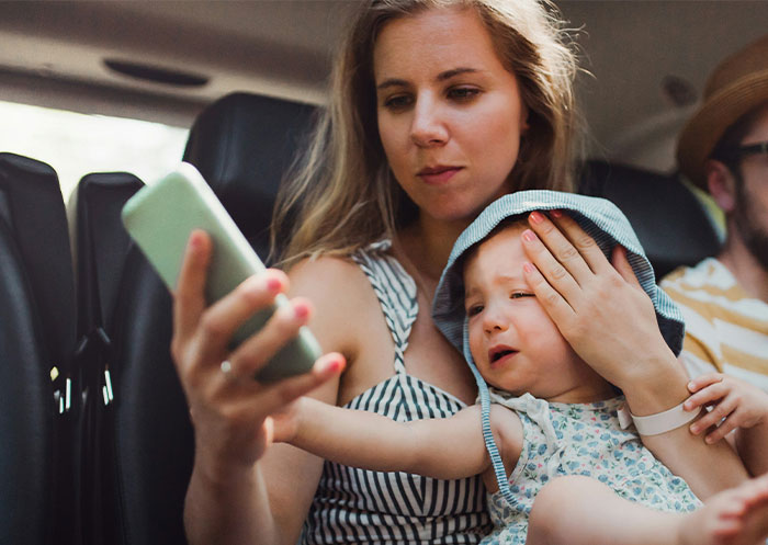Woman ignores upset child while looking at phone during family vacation with man-child husband in backseat of car.
