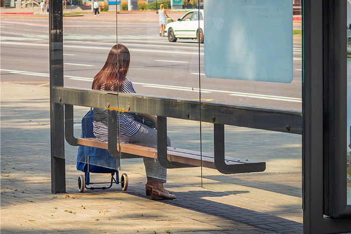 Woman sitting at a bus stop with a shopping trolley, waiting on a sunny day near a busy street in Boston marathon area. - 19
