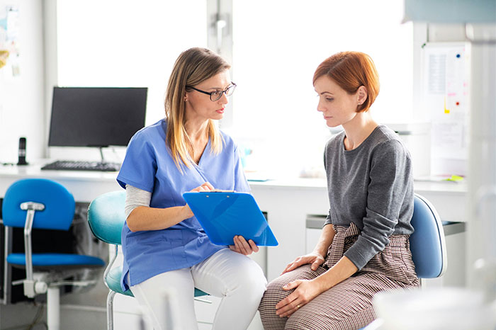 A nurse in blue scrubs discussing health details with a female patient in a bright medical office setting. - 34