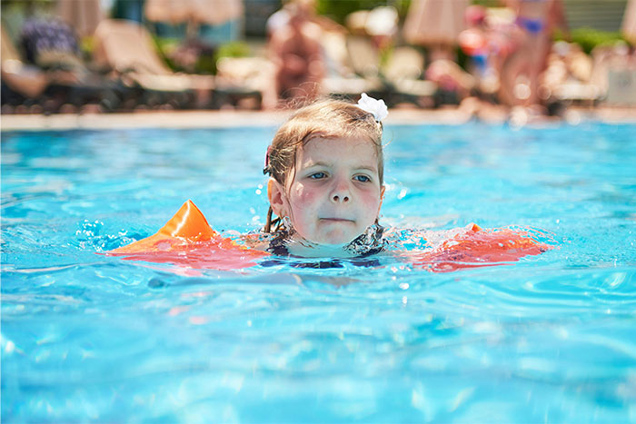 Young child wearing orange floaties swimming in a pool on a sunny day with people blurred in the background. - 10