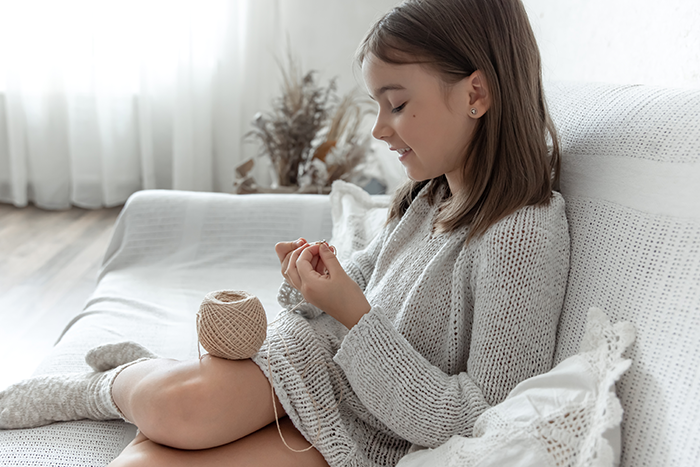 Young girl knitting with yarn while sitting on a white sofa in a cozy living room setting.