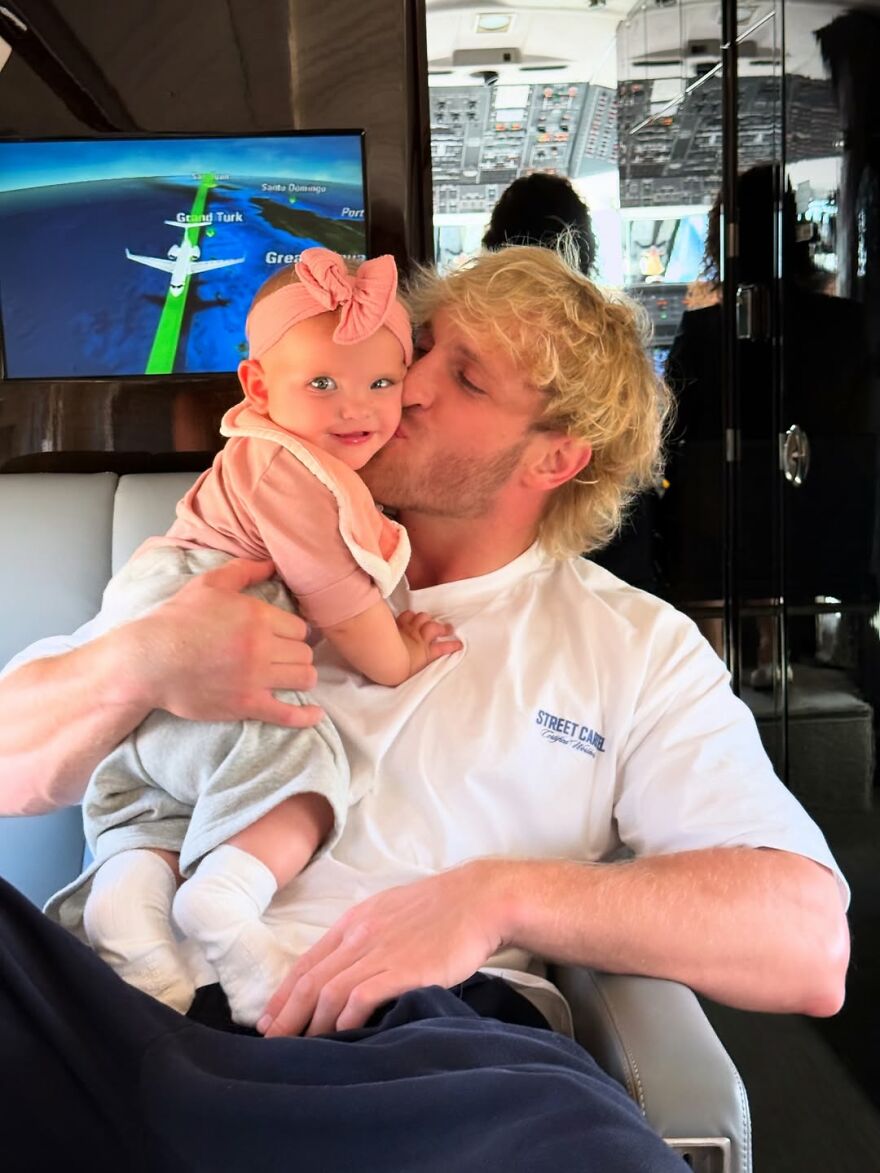 Jake Paul holding and kissing a baby inside an airplane cabin with a flight map screen in the background.