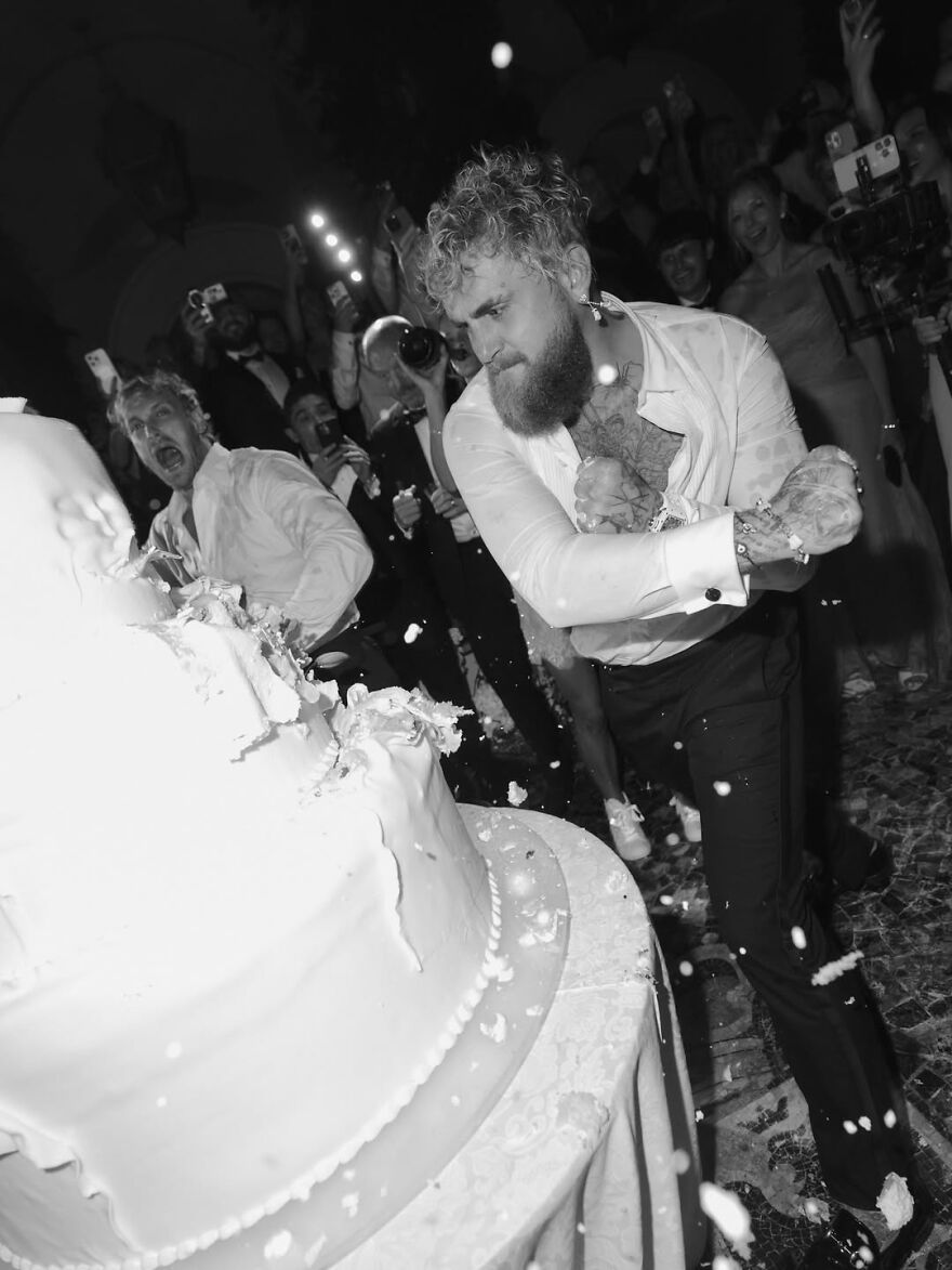 Jake Paul smashing wedding cake at event with crowd reacting and capturing the bizarre viral moment in black and white.