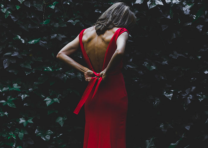 Woman in a red dress tying a bow in a dark outdoor setting surrounded by leaves, evoking eerie road encounter themes.