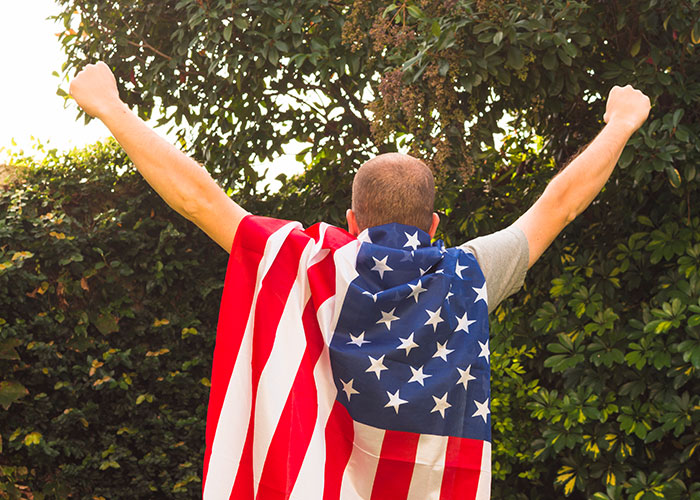Man wrapped in an American flag raising arms outdoors, symbolizing strength and resilience of truck drivers on the road.