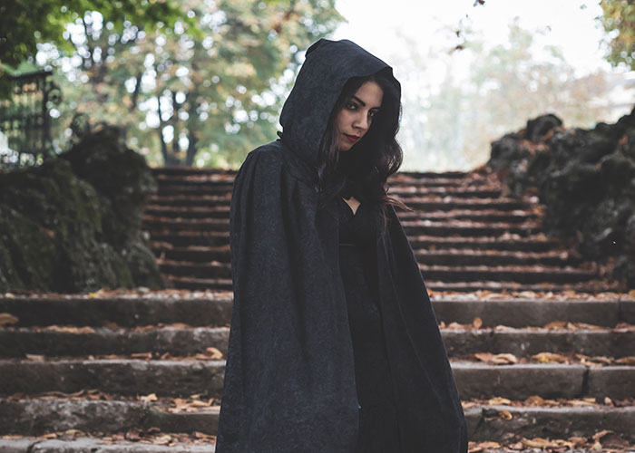 Woman in dark hooded cloak standing on weathered stone steps in eerie outdoor setting, evoking terrifying road encounters.