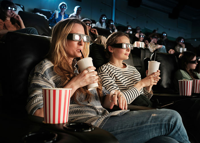 Two women wearing 3D glasses and drinking soda at a movie theater, enjoying a relaxed entertainment experience.