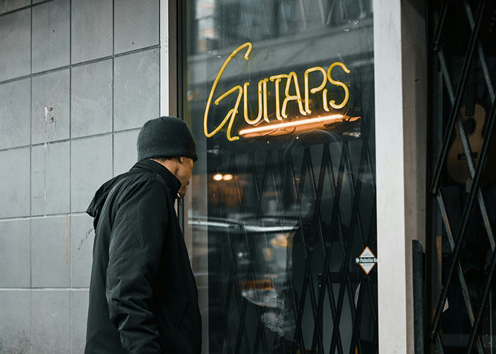 Man wearing a black beanie and jacket looking through a glass door at a neon sign in a store displaying guitars loopholes.