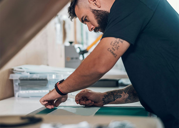 Man with tattoos and a beard leaning over a table, focused on a project using a tool in a workshop setting.