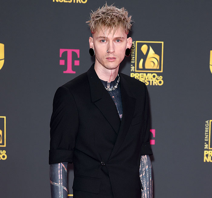 MGK in a black suit with tattoos visible on arms and neck, attending a music awards event on the red carpet