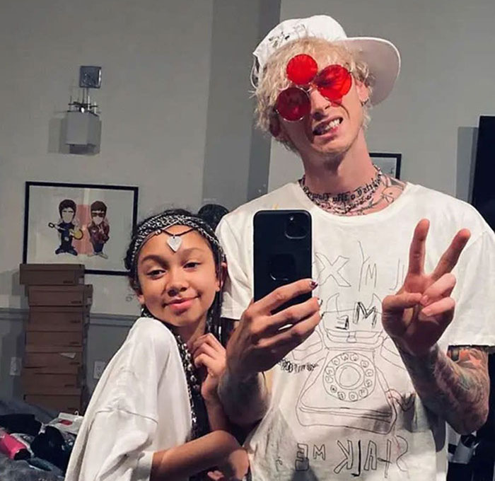 MGK wearing red sunglasses and white cap takes selfie with a young girl, reflecting his half alien claim in casual setting.