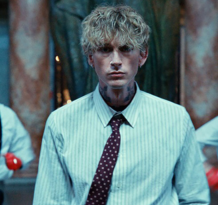 MGK with blonde curly hair and tattooed neck, dressed in shirt and tie, related to claim of being half alien in interview.