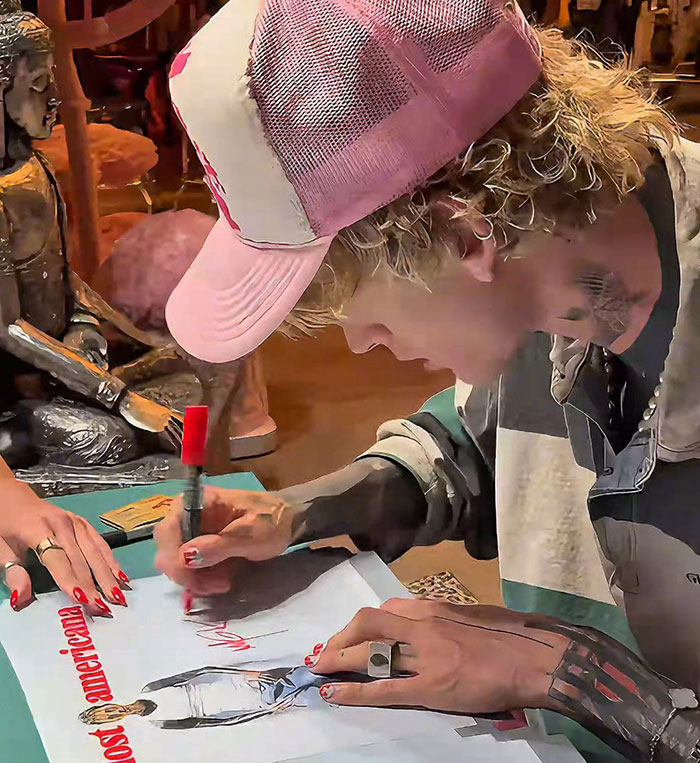 MGK wearing a pink cap and signing artwork, highlighting claims about being half alien and age uncertainty in an interview.