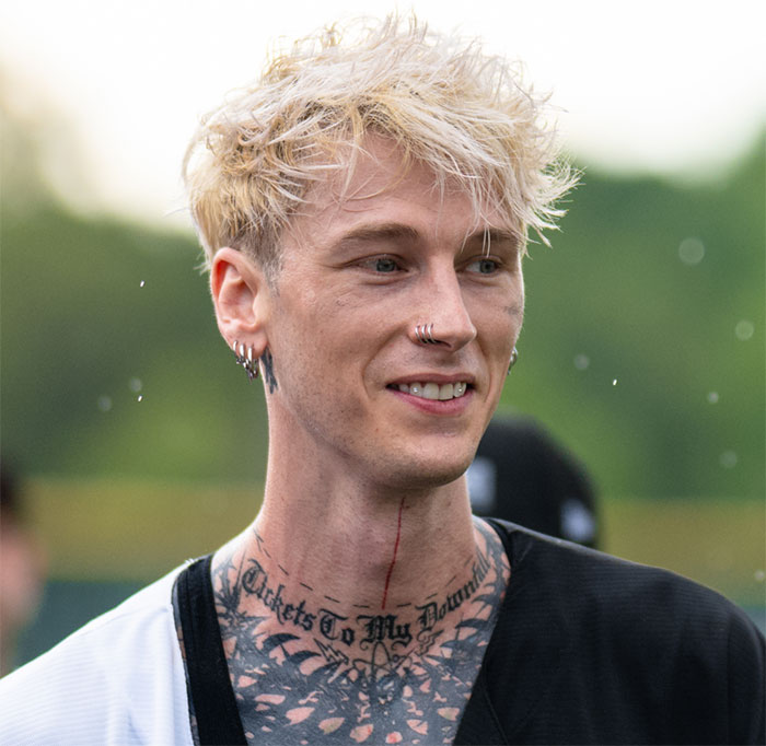 MGK with platinum blonde hair and tattoos, smiling outdoors during an interview about his alien claims and age mystery.