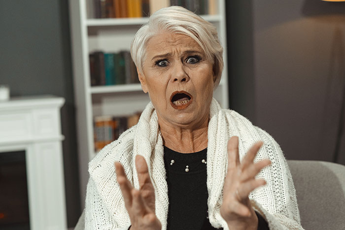 Older woman with short white hair looks shocked and upset while gesturing with her hands in a home setting