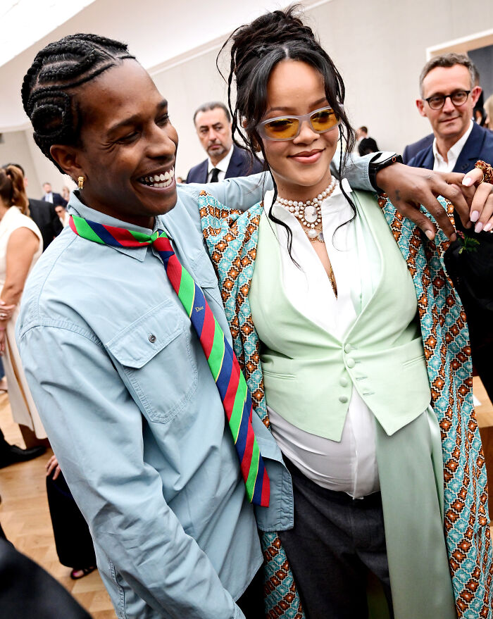 Rihanna wearing maternity dress with patterned coat and sunglasses, smiling with man in blue shirt and colorful tie.