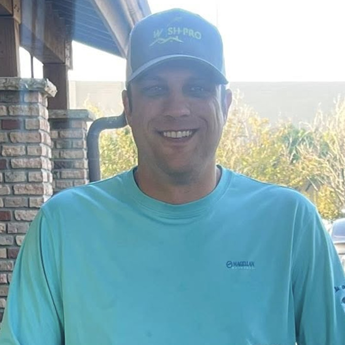 Man wearing a blue shirt and cap, smiling outdoors with sunlight and brick wall in the background, related to breast milk assault case.