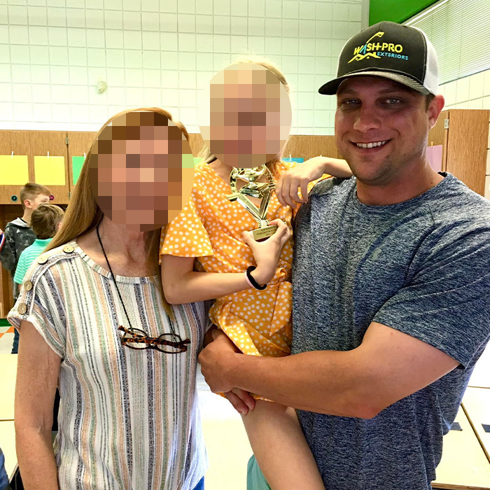 Man wearing a black cap holding a child in a yellow dress with an adult woman nearby in a casual indoor setting.