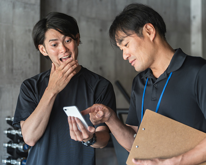 Man surprised looking at phone while gym staff points, discussing expensive gym membership issue indoors. Man surprised looking at phone while gym staff points, discussing expensive gym membership issue indoors.
