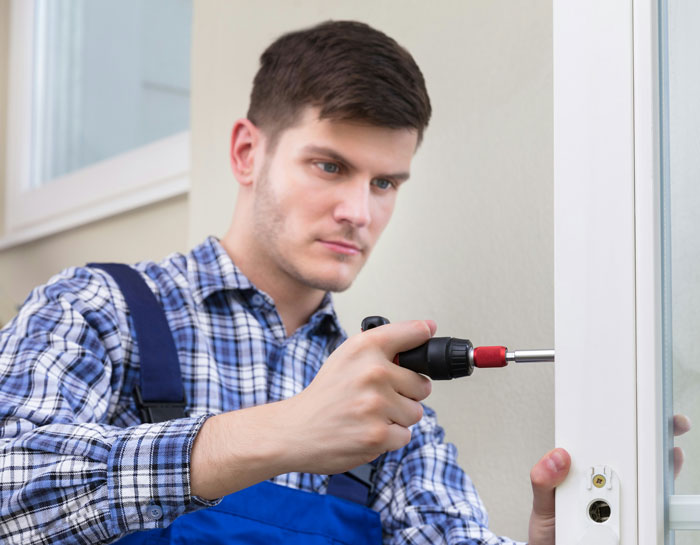 Handyman in blue overalls installing new cat flap with electric screwdriver on white door frame inside home.