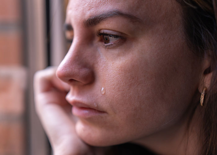 Sad woman with tear on cheek looking out window, reflecting on boyfriend excluding her from couples trip without asking.
