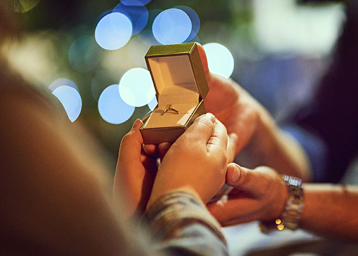 Hands holding an open ring box with an engagement ring, representing a woman discovering boyfriend told friends she couldn&rsquo;t join trip.