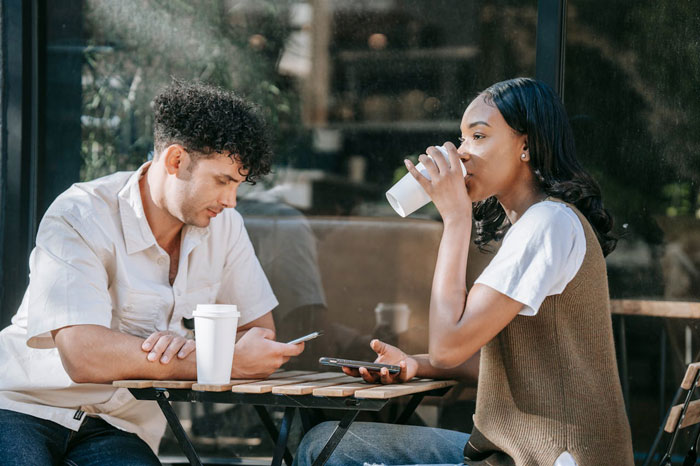 Young woman and man sitting at an outdoor cafe, woman drinking coffee while man looks at his phone, depicting friend&rsquo;s bf as ATM.