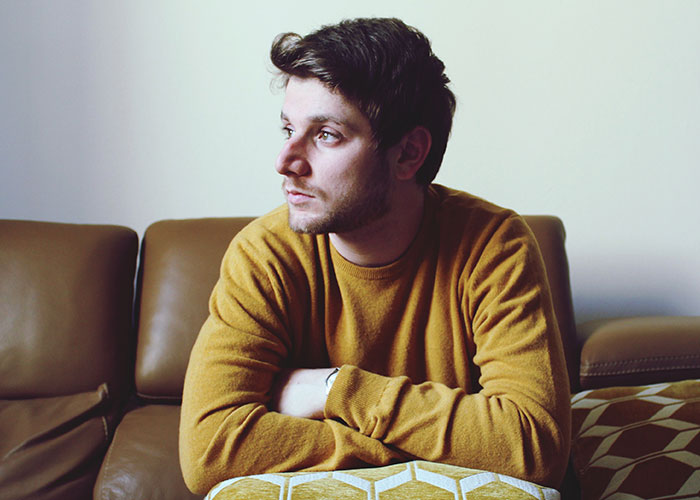 Young man in a mustard sweater sitting on a sofa, looking away thoughtfully, relating to tattoo and late son themes.