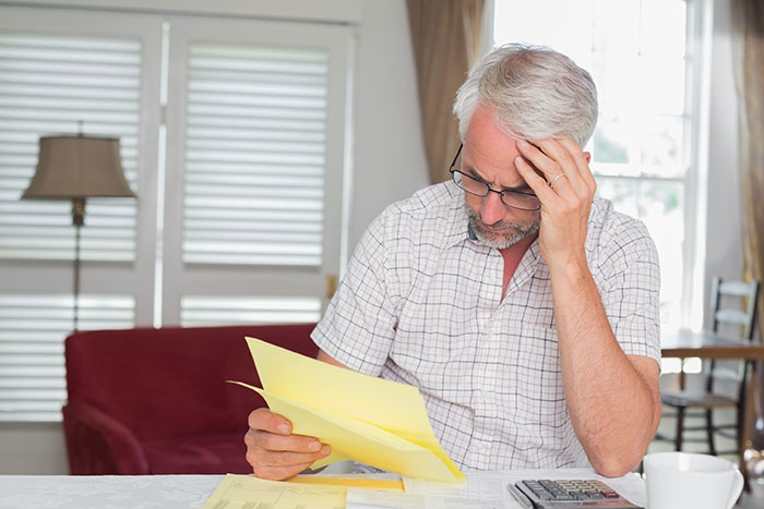 Man with gray hair and glasses looking stressed while reviewing documents at home, representing wedding expense concerns.