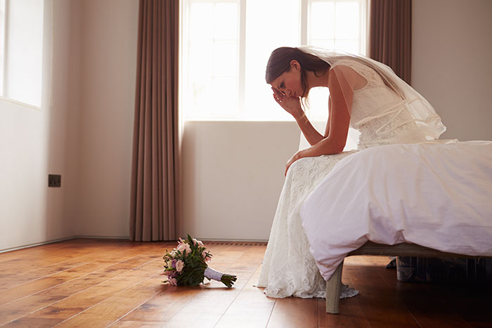 Bride sitting on bed looking upset with wedding bouquet on floor, reflecting a failed wedding moment.
