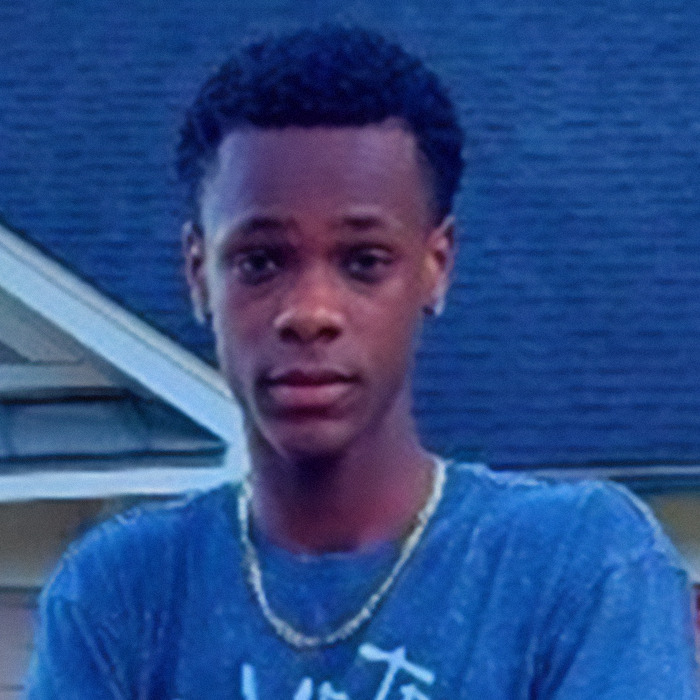 Young man wearing a blue shirt and gold chain standing outside a house, related to Batman villain and homicide news.