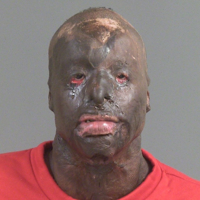 Man resembling Batman villain with extensive facial tattoos and scars wearing a red shirt posing for mugshot photo.
