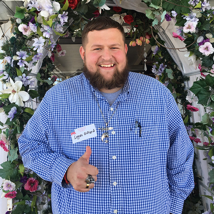 Man with beard and checkered shirt smiling and giving thumbs up standing under floral arch in bright setting