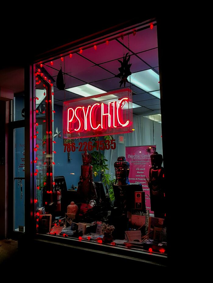 Neon psychic sign glowing in a storefront window decorated with statues and red string lights at night.
