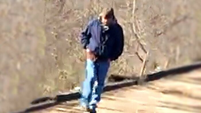 A man in a dark jacket and blue jeans walking on a path with bare trees in the background, context unclear. - 21