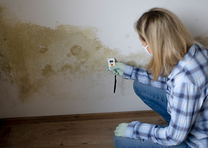 Woman wearing mask and gloves using a device to check mold on a wall, highlighting risks of patients being too young to be sick.