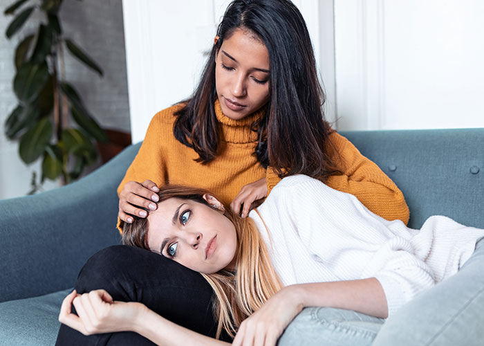Young woman comforting sick friend on couch, illustrating doctors getting it wrong and patients suffering consequences.